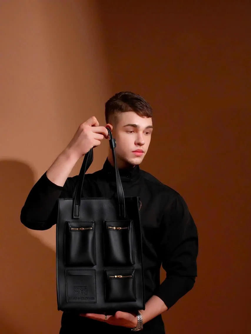 A model in black lifts the DK 01 Moscow tote by its handles against a warm brown studio wall, showing the grid of front pockets.