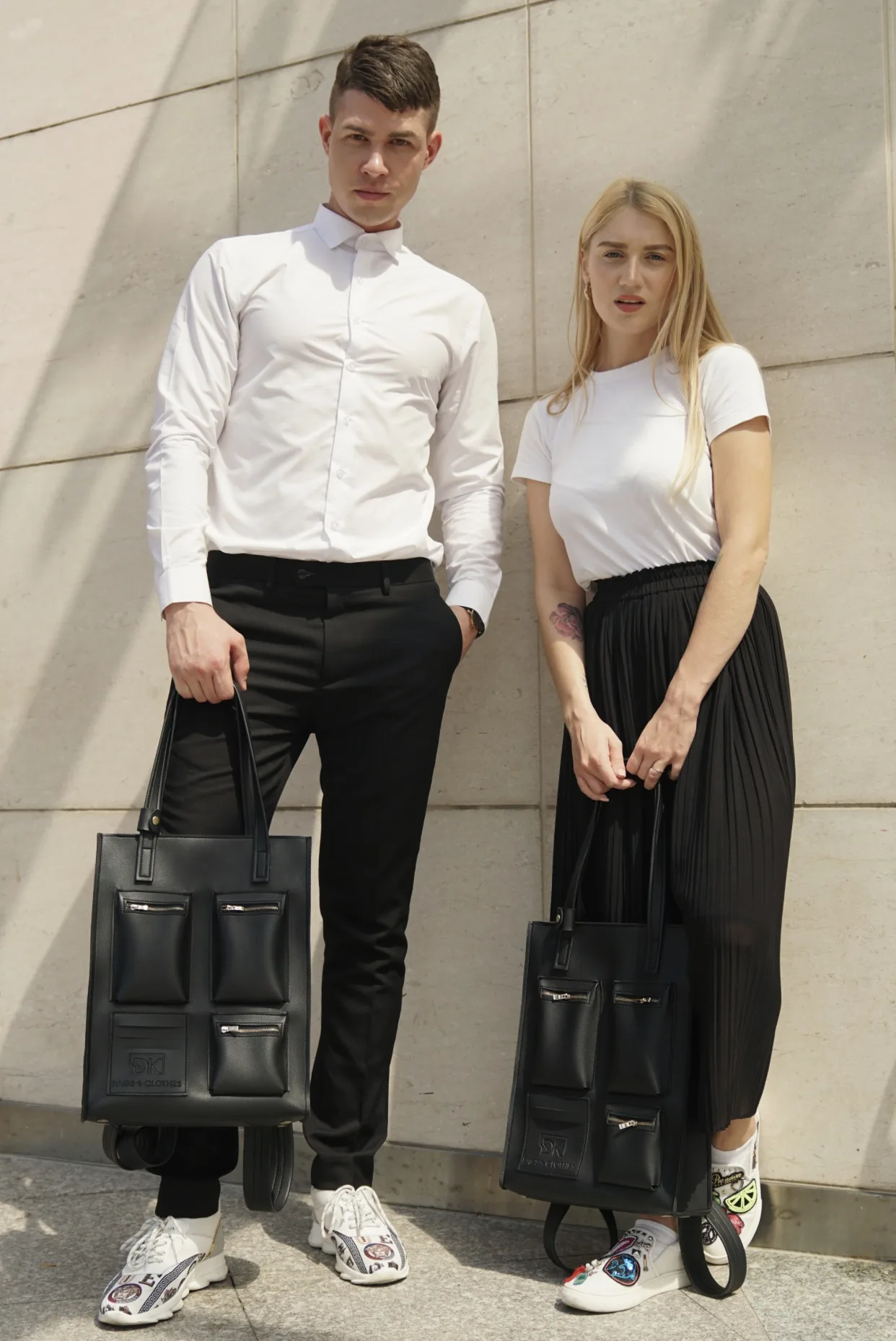 Two models in white shirts and black trousers stand against a travertine wall, each holding a black DK 01 Moscow tote by its handles.